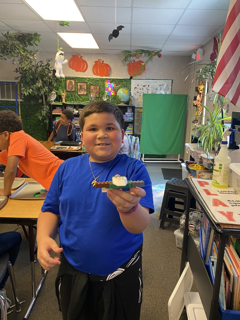 A young boy in a blue shirt and black pants displays a small, edible craft. Behind him, a classroom is decorated with fall themes.