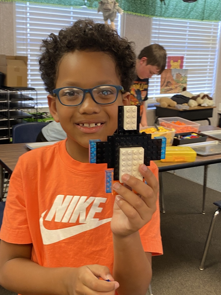 A child wearing glasses and an orange shirt holds a Lego structure in a classroom.
