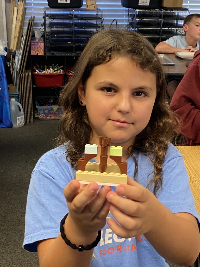 Girl in a blue shirt holds up a small Lego bridge. Behind her, a blurred person sits at a table.