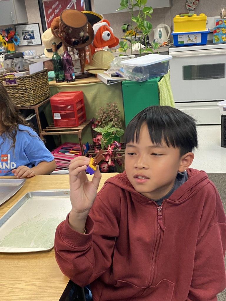 A child in a maroon hoodie examines a small object, seated at a table. A girl in a blue shirt sits behind him.