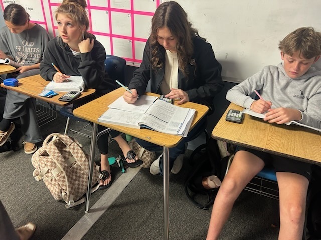 A classroom with students seated at desks, some with backpacks, all engaged in writing on paper.