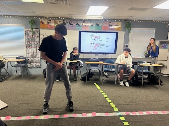 A classroom with desks, a teacher, and a student practicing a performance. Bright lighting, a screen, and floor markings.
