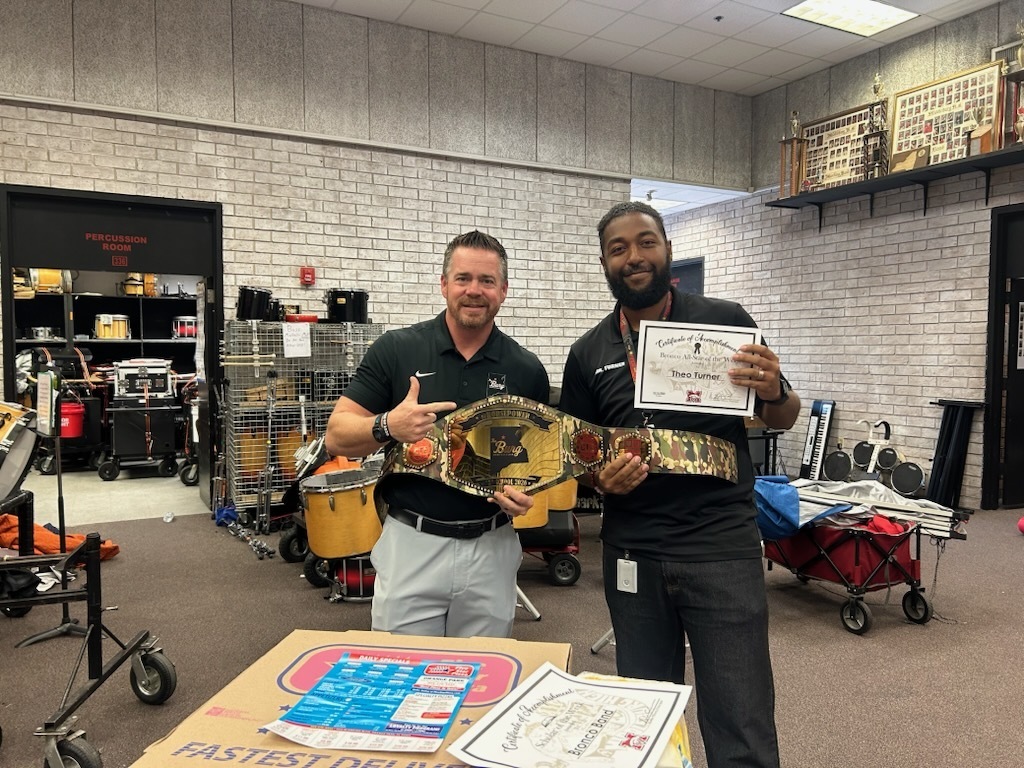 Two men stand with a championship belt and a certificate in a room with musical equipment.
