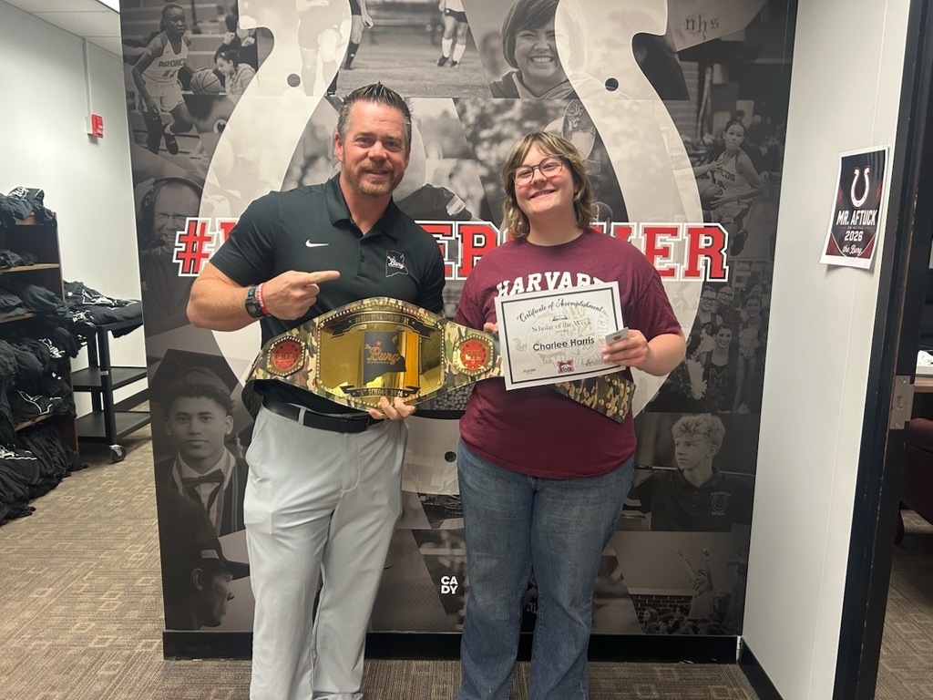 Two people pose in front of a sports-themed wall, one holding a championship belt, the other holding a certificate.