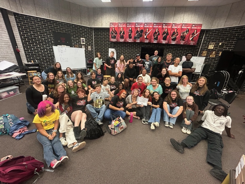 A group of individuals sit on the floor in a room with brick walls, one holding a guitar.
