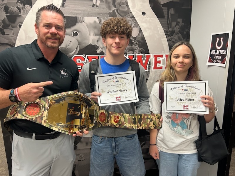 Three people, one man and two women, stand next to each other, holding certificates and a wrestling belt.