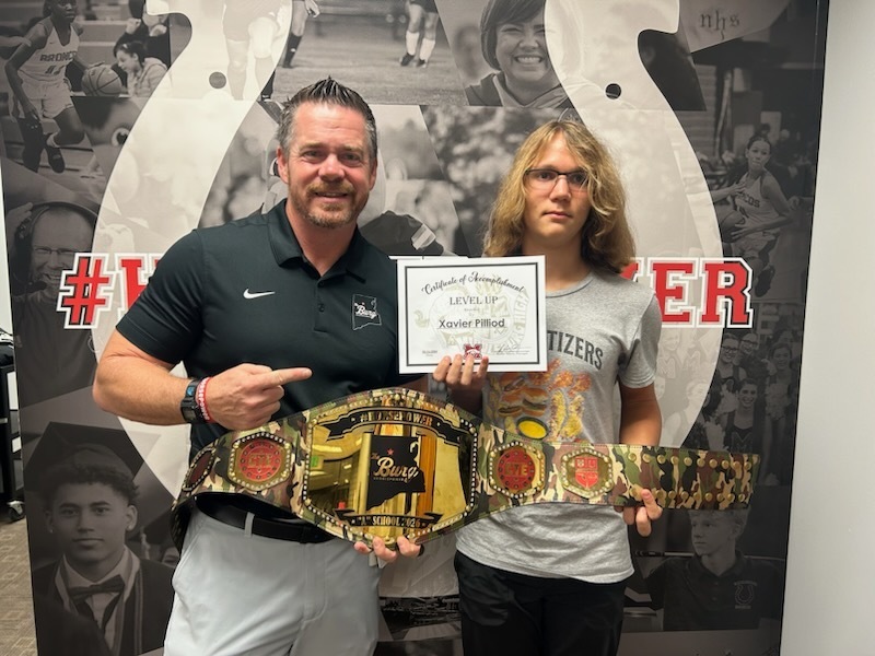 Two people, a man and a woman, stand in front of a photo wall, displaying a championship belt and certificate.