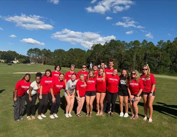 Group of people in red shirts and shorts standing on a golf course, trees in background.