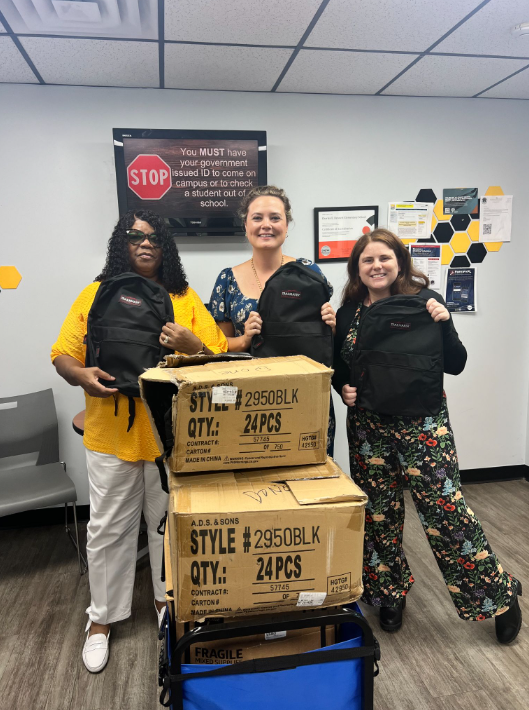 Three women standing behind a cart with boxes, each holding a backpack. Boxes labeled with quantity and style.
