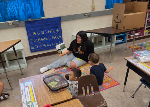 Woman sitting on a rug with two children, reading a book, in a classroom with desks and chairs.