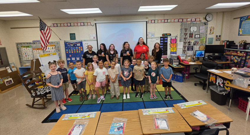 Children and adults stand in a classroom with desks and a rug. A flag and bulletin board are present.