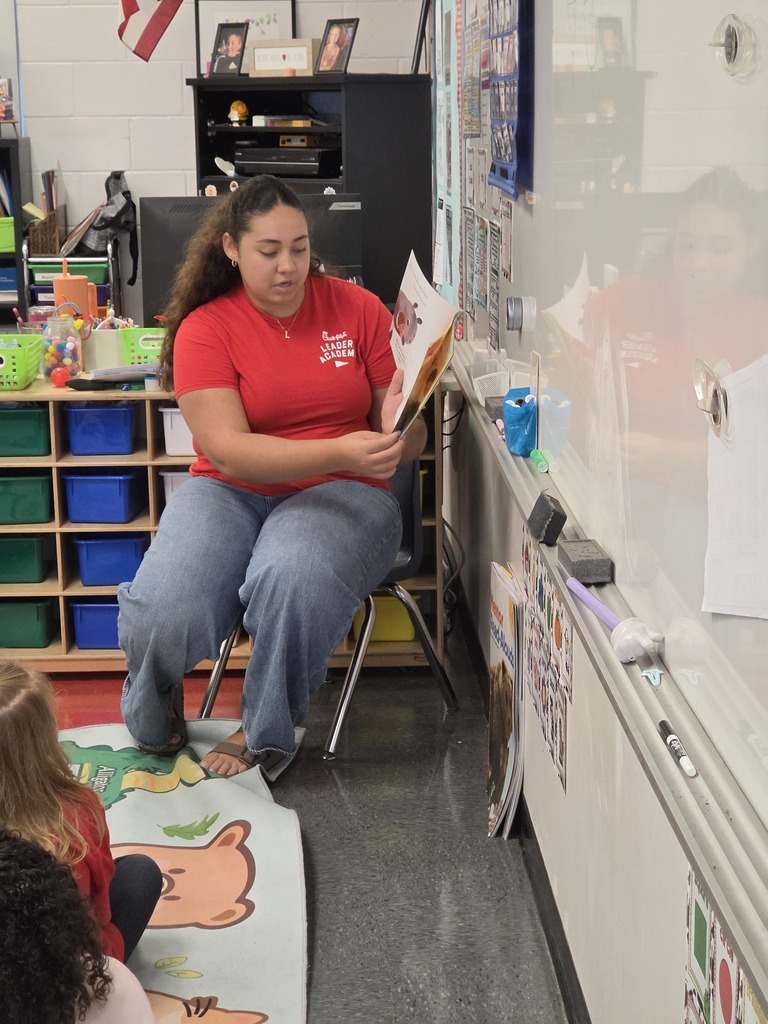 A woman in a red shirt and jeans reads aloud from a book to children sitting on the floor.