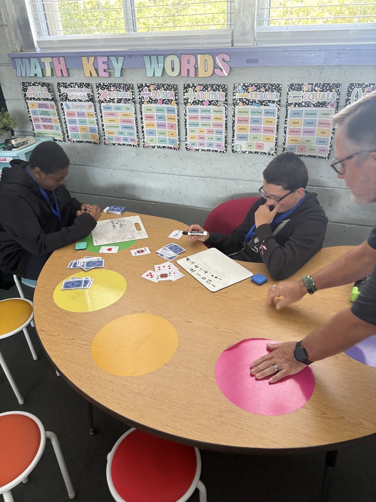 Three people sit at a table with multicolored circles, one handling cards and another using a marker.