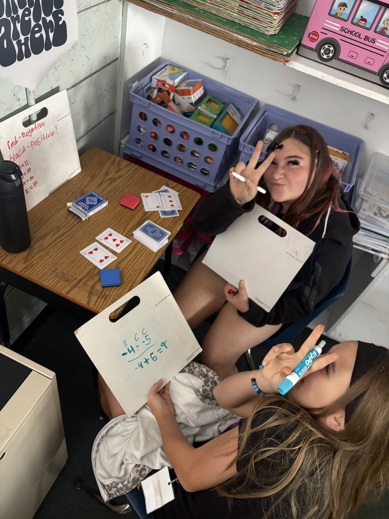 Two students seated at a desk, one holding a marker and writing on paper, the other holding a bag.