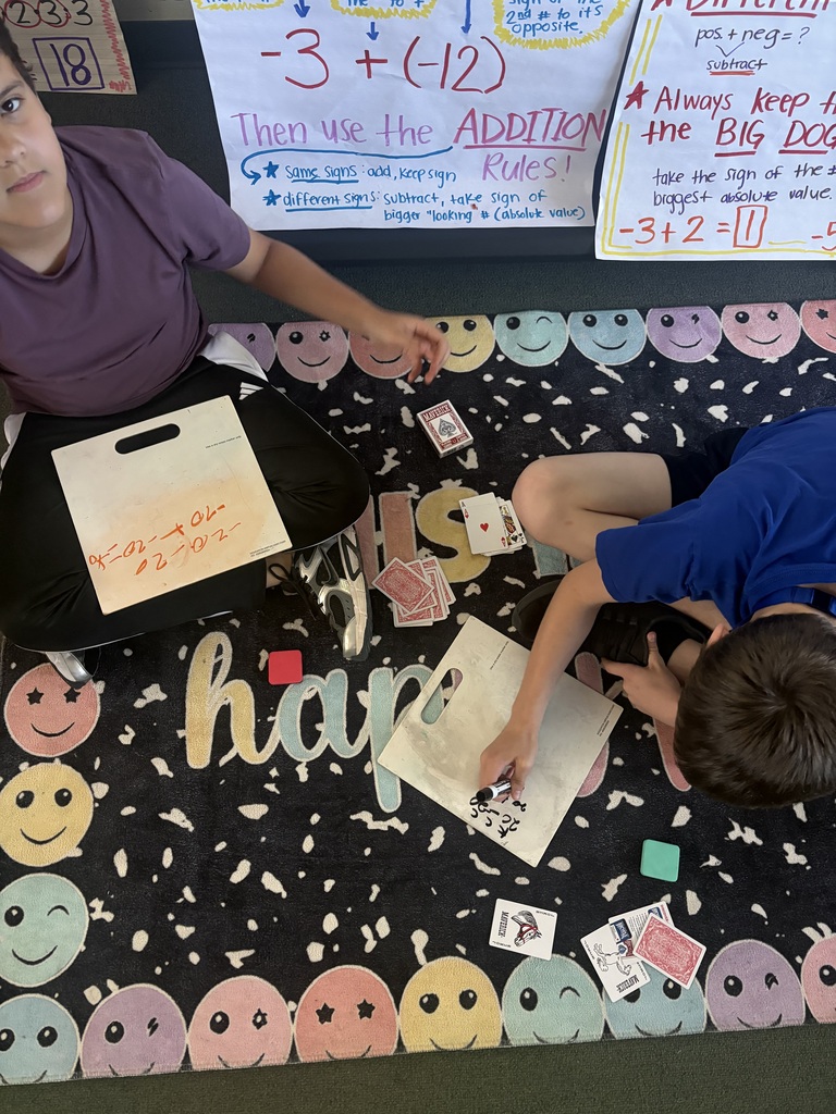 Two children seated on floor, working on math problems with cards, in front of posters.