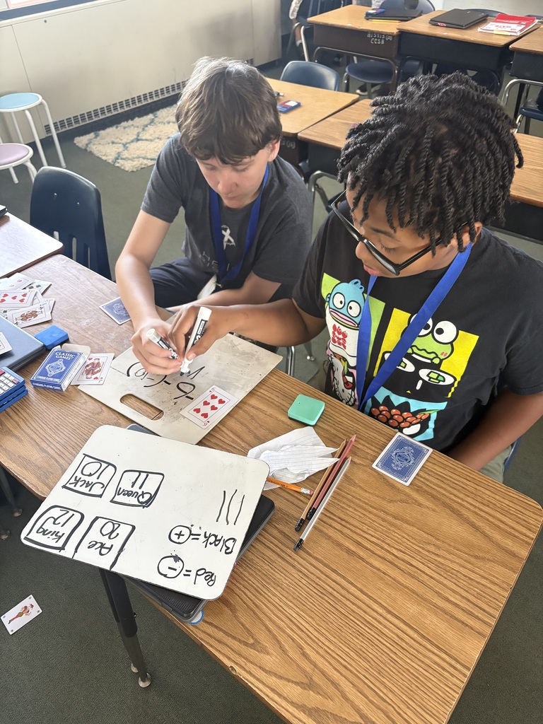 Two students in a classroom work on a table with papers, markers, and a whiteboard.