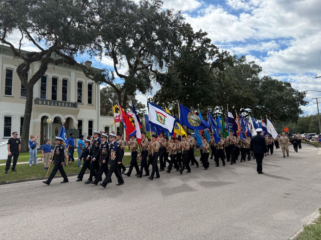 Marching group carrying flags in front of building with people walking on the sidewalk.