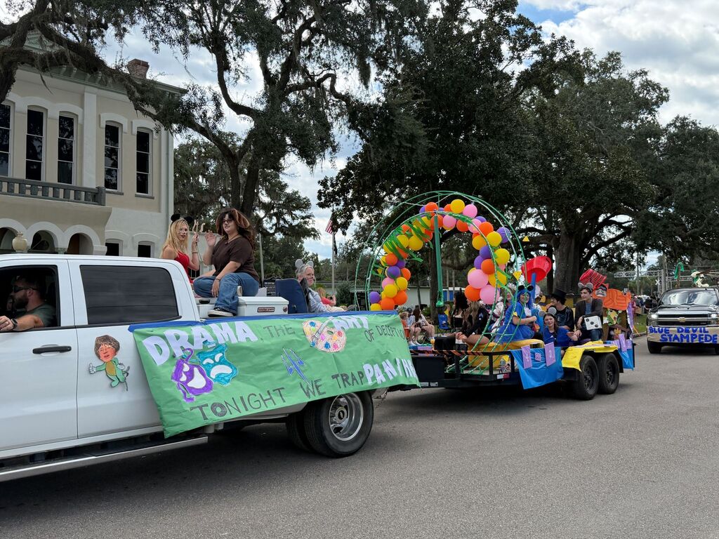White truck and trailer with green banner with "Derma Tonight at the Park" float behind, passing by trees and buildings.