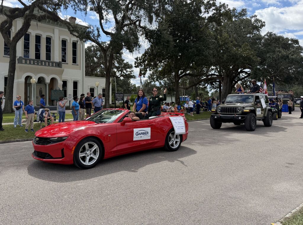 Red convertible car with two people in it and white banner on front, with SUV behind.
