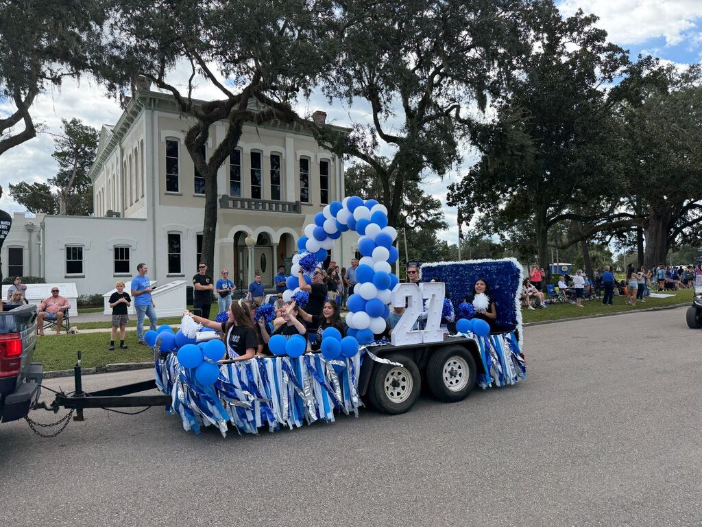 Parade float with blue and white decorations, including balloons and a number 21, moving past a building.