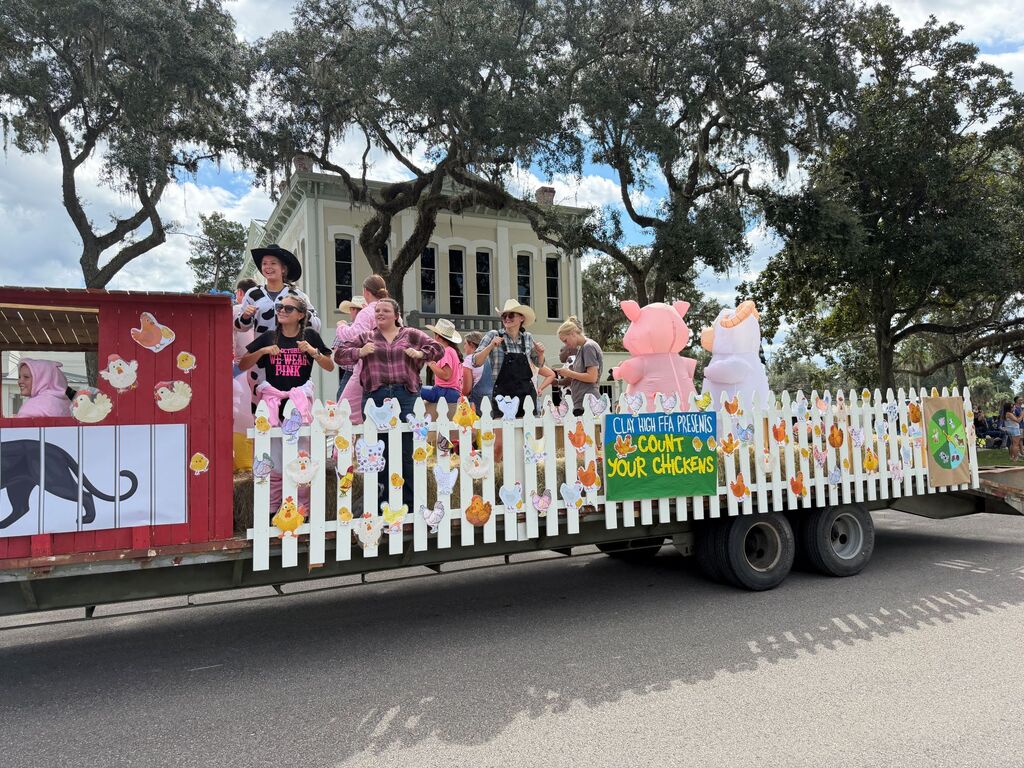 Parade float with a white picket fence in front. Two pink pigs, two adults, and three children.