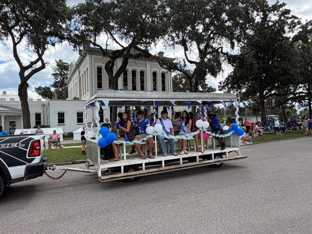 People sit on a white and blue parade float, some holding balloons, as it is towed by a truck.