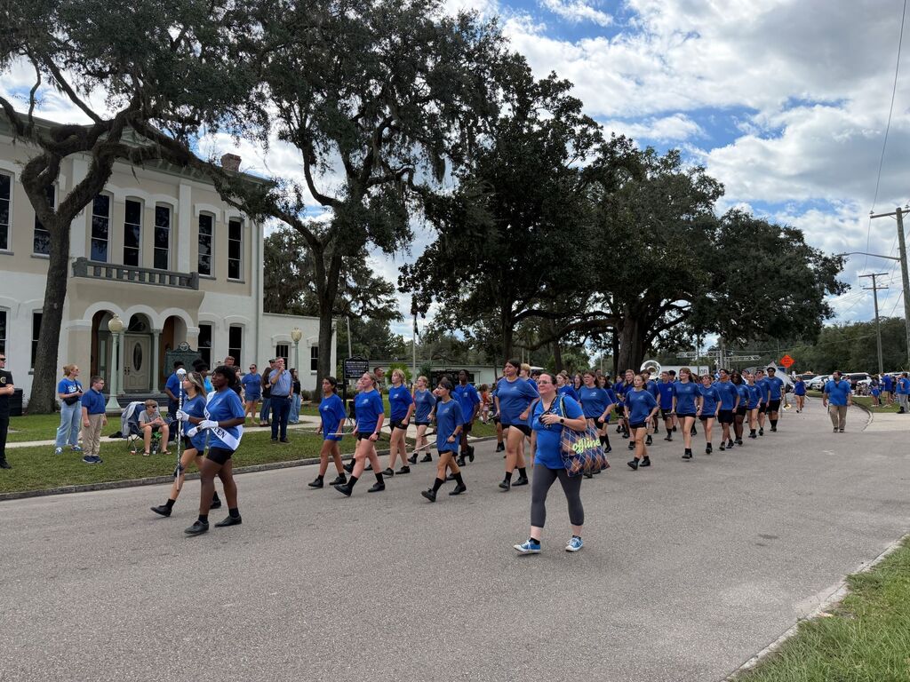 Group of people in blue shirts and shorts running in a line on a road with trees.
