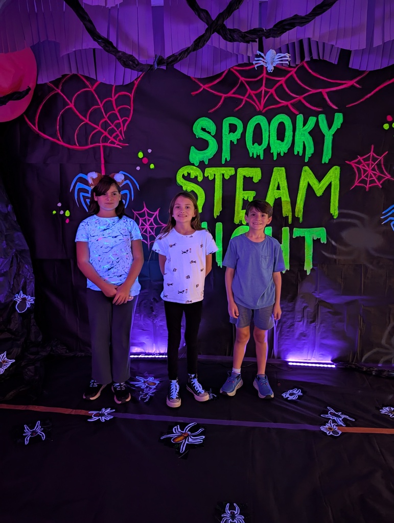 Three children stand on a stage in front of a backdrop with glowing purple and green letters.