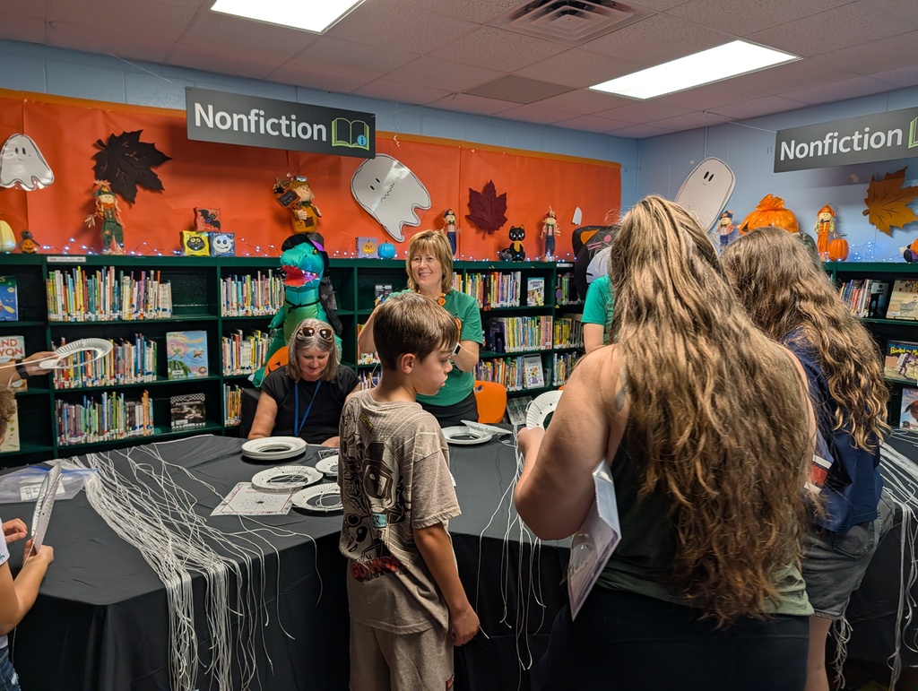 Library with shelves of books. A group of children and adults stand around a table with paper.