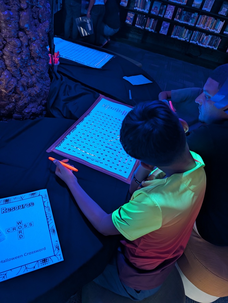 Two children seated at a table with papers. One is holding a pen and pointing to a crossword puzzle.