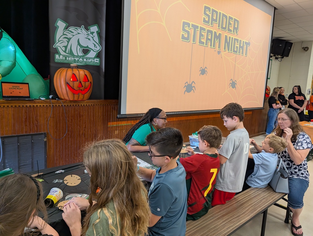 Students sitting at tables for a STEAM event, with a pumpkin and screen showing "Spider STEAM Night" in the background.