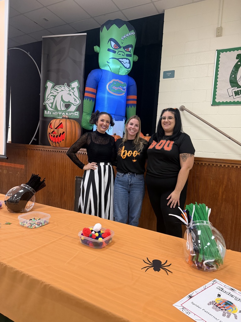 Three women standing behind a table with orange cloth and decorative items. A large green inflatable is in the background.
