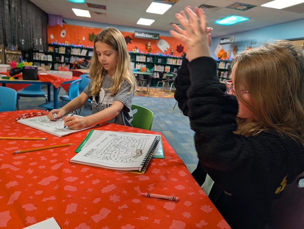 Two girls in a classroom with books and pencils. One is writing and the other is raising her hand.
