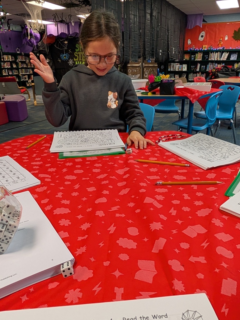 Child seated at a table with red tablecloth, using pencil and paper, in a classroom with bookshelves and chairs.