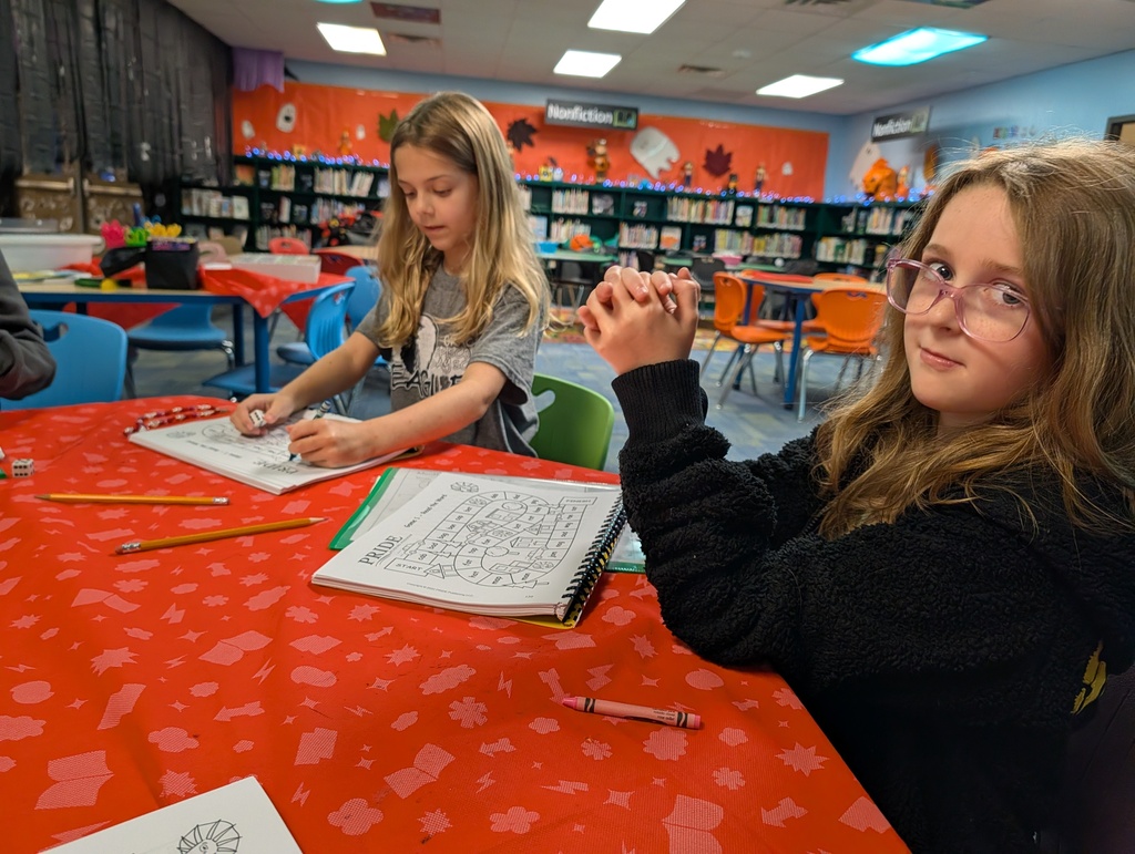 Two girls seated at a table, one holding a pen, writing in a notebook. One girl wearing glasses.