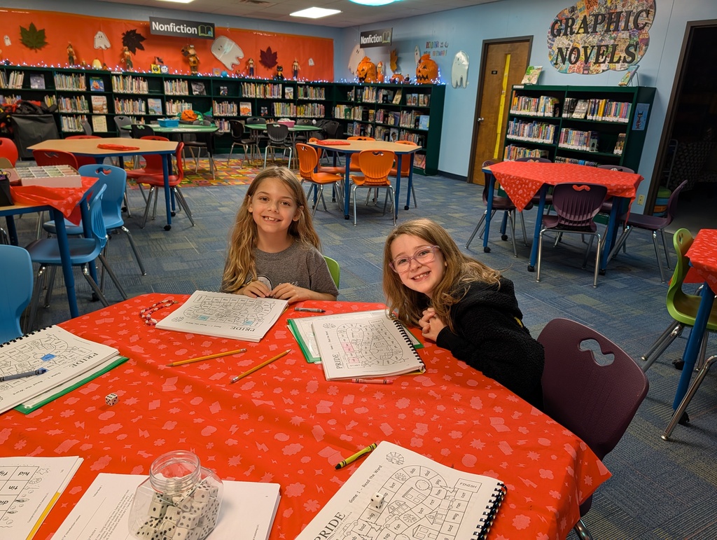Two young girls sitting at a round table in a library, smiling, each holding a paper and pencils.