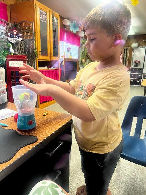 Boy in yellow shirt stands in classroom, holding a toy blender and turning it on table.