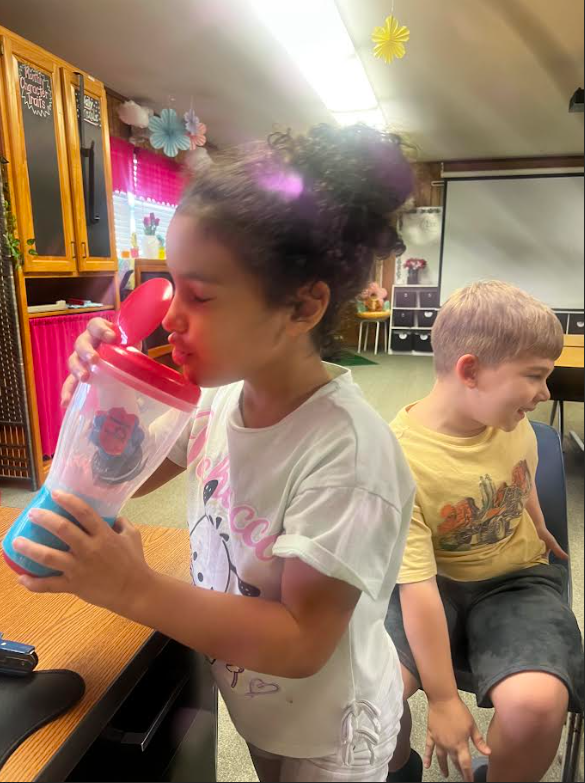 Girl holding blue cup with red lid up to her face, seated boy wearing yellow shirt with black shorts nearby.