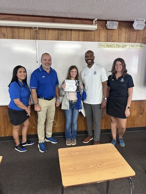Five people in a classroom, four in blue and one in white, pose with a young person holding a certificate.
