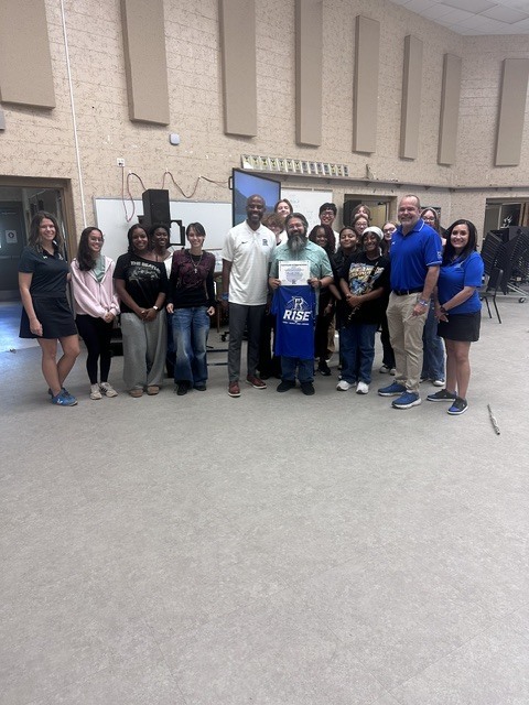 A group of adults and students pose for a photo in a room with a stage and a whiteboard.
