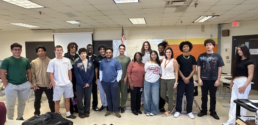 Students and staff stand in a classroom with a projector screen and American flag in the background.