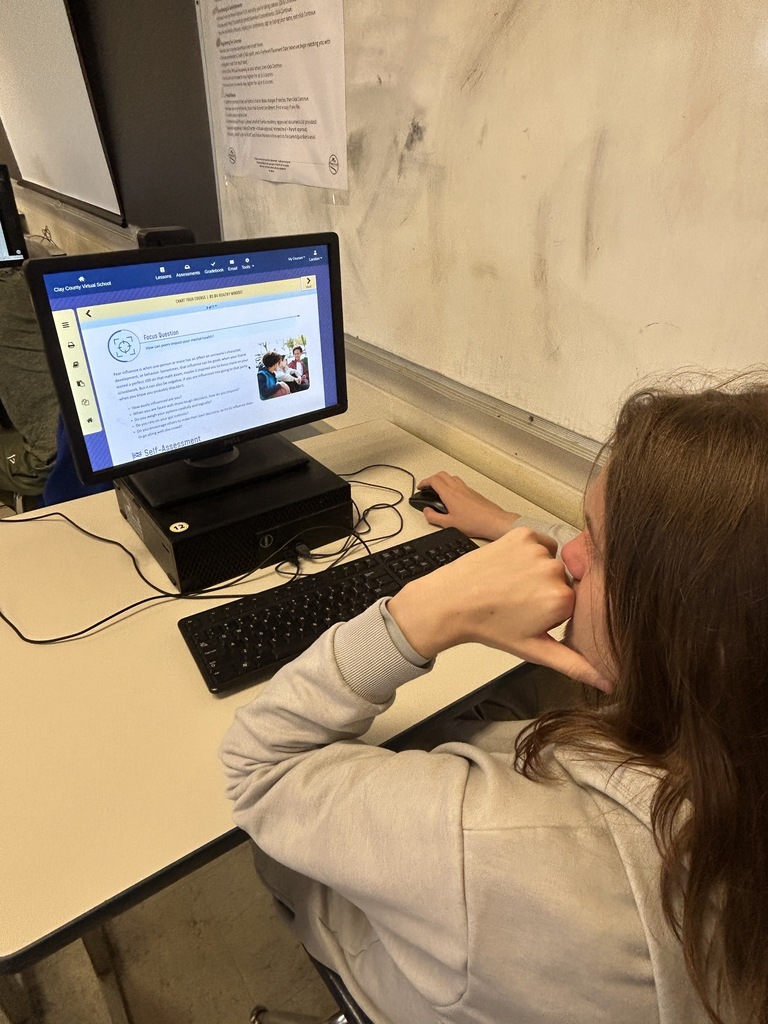 A student sits at a desk using a computer. A white board is behind them.