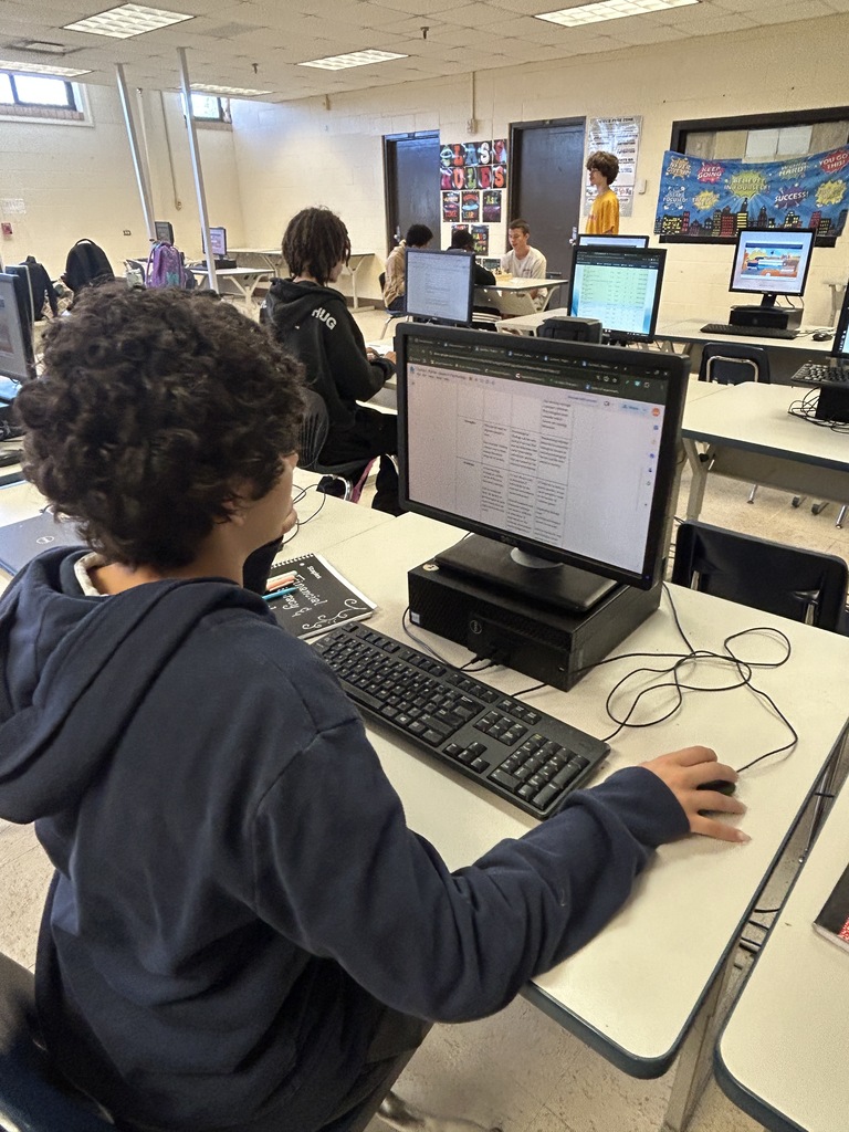 A student sits at a computer in a classroom, typing on a keyboard, with other students in the background.