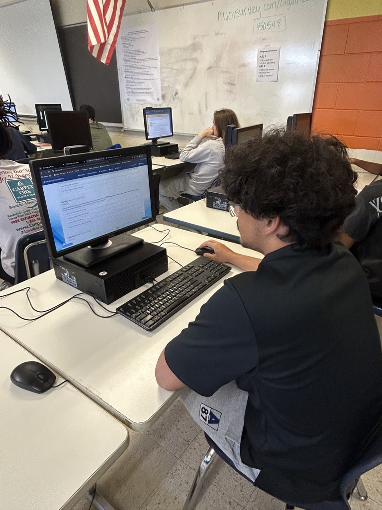 Student sits at a computer on a desk in a classroom. Two other people work behind him.