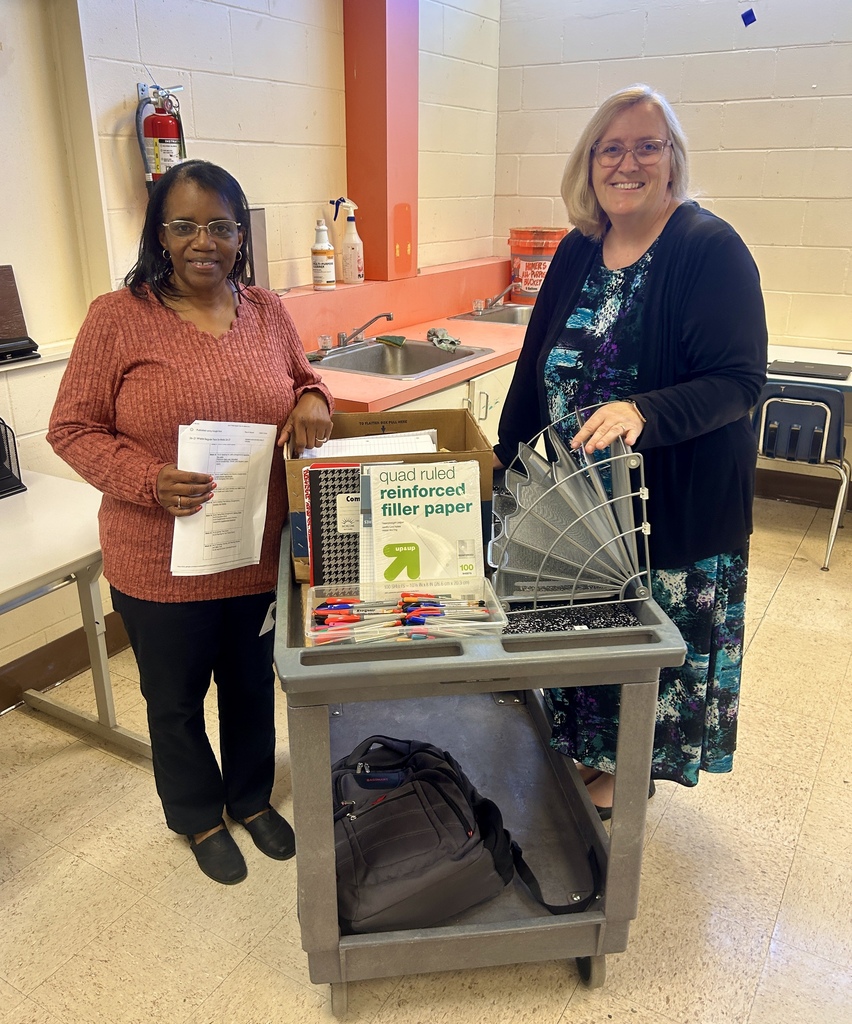 Two women standing next to a cart with school supplies, one holding a paper and the other holding a box.