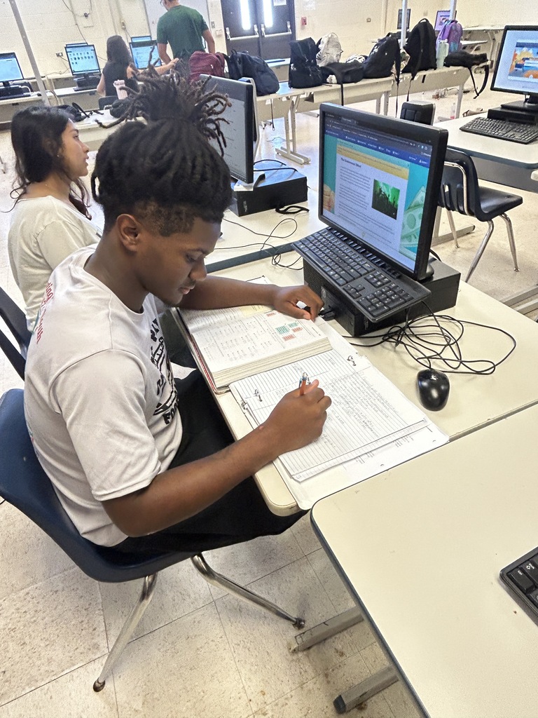 Students seated at tables in a classroom, with one writing in a book and a computer nearby.