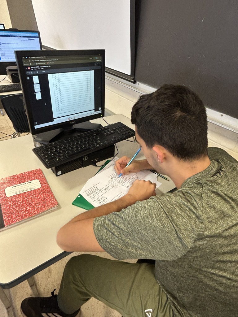 A student writes on paper at a desk in front of a computer monitor.