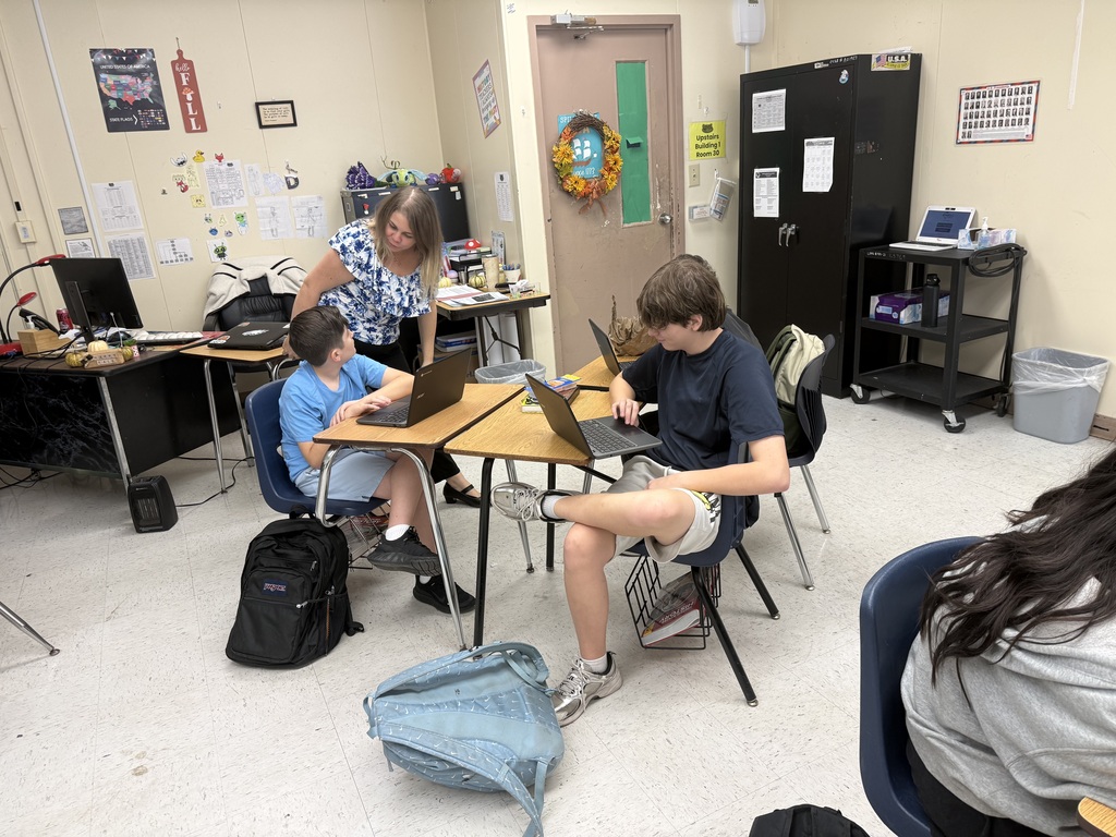 Three students sit at desks with laptops, one with legs up. A woman stands behind a desk. A trash bin stands in the background.