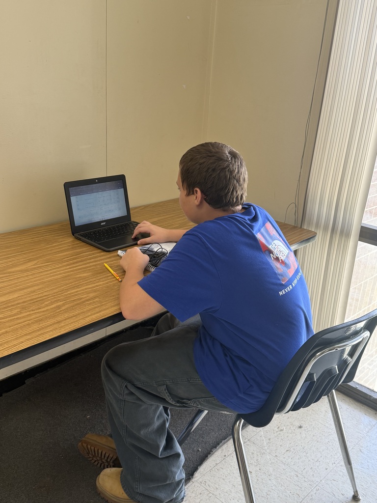 Student seated in chair at desk, typing on laptop; pencils and papers on table; glass windows behind student.