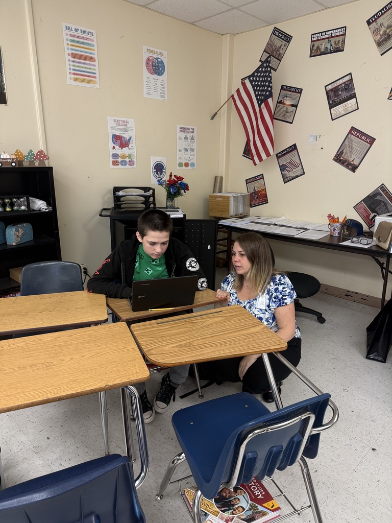 A student uses a laptop while sitting at a desk in a classroom. A teacher kneels beside him.